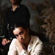 Woman in a white outfit with black patterns sitting in a natural setting with a blurred man in the background.