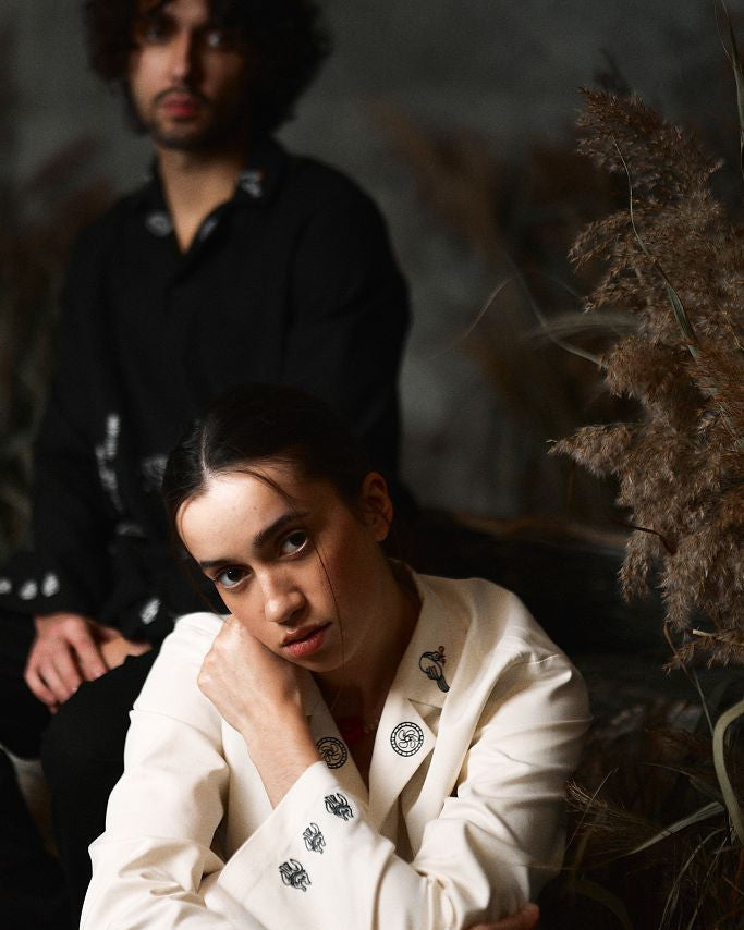 Woman in a white outfit with black patterns sitting in a natural setting with a blurred man in the background.