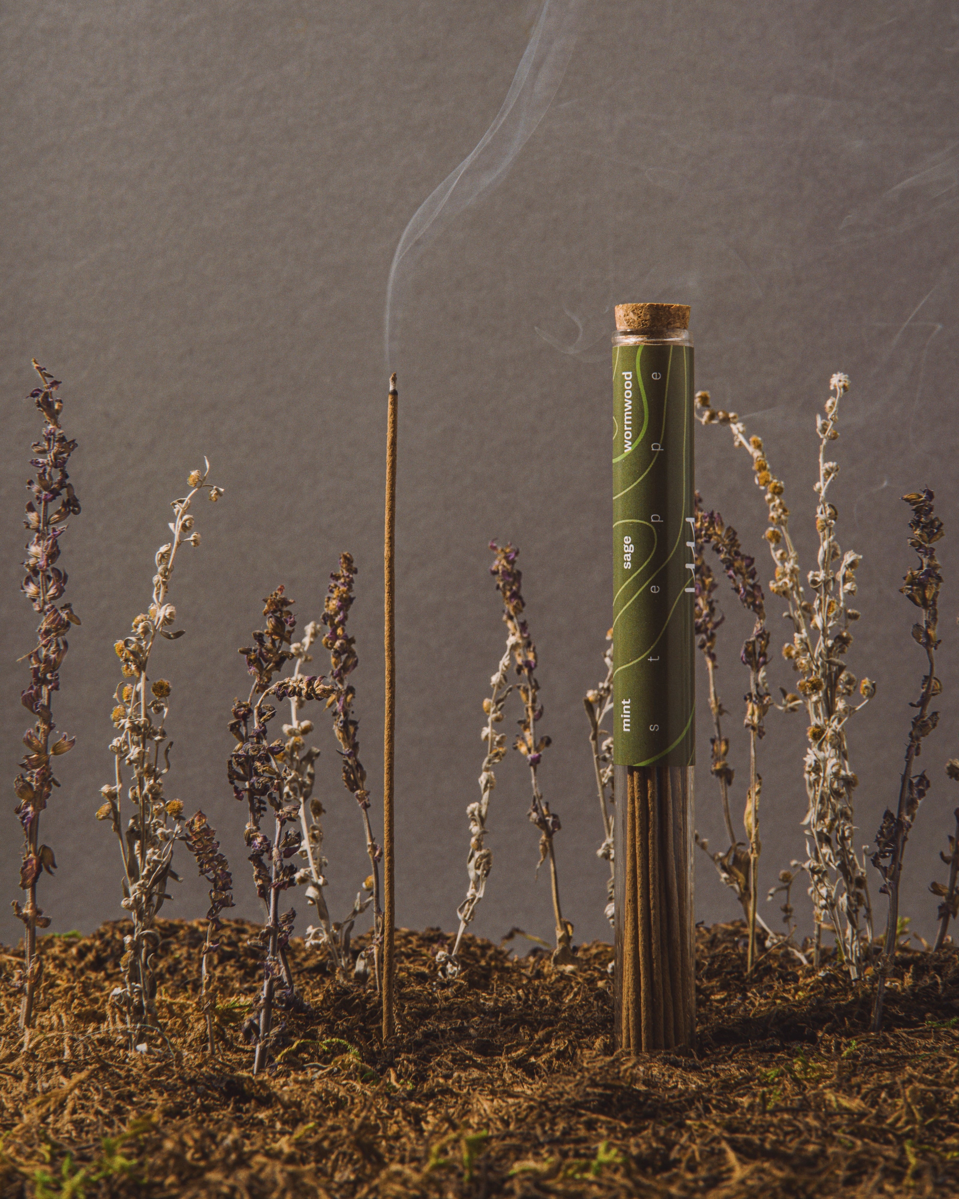 Cigarette with a lit end surrounded by small plants on a gray background