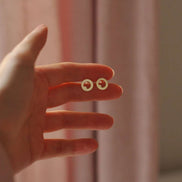 A hand holding a pair of small gold hoop earrings with tiny red circular details inside, set against a soft pink curtain background.