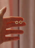 A hand holding a pair of small gold hoop earrings with tiny red circular details inside, set against a soft pink curtain background.