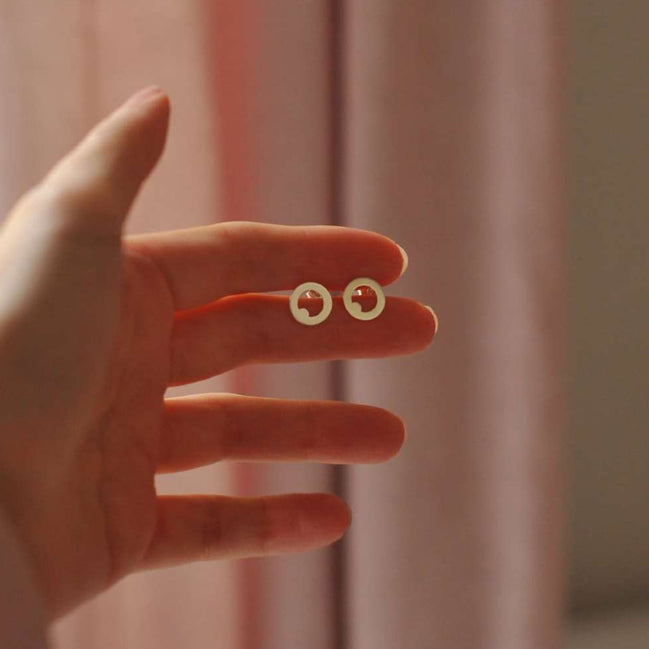 A hand holding a pair of small gold hoop earrings with tiny red circular details inside, set against a soft pink curtain background.