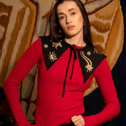 Woman wearing a red top with black embroidered collar against a patterned wall.