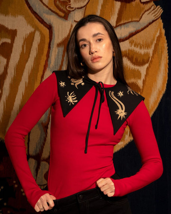Woman wearing a red top with black embroidered collar against a patterned wall.