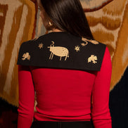 Person wearing a red top with a black shoulder cape featuring gold embroidery against a patterned wall.