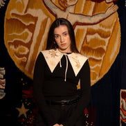 Woman in a black outfit with white collar standing in front of a decorative tapestry.