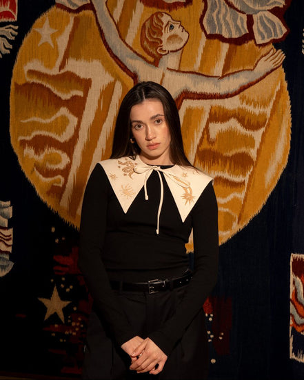 Woman in a black outfit with white collar standing in front of a decorative tapestry.