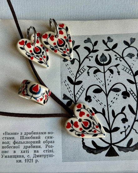 Heart-shaped ceramic earrings with red and black patterns on a piece of paper with black line art.