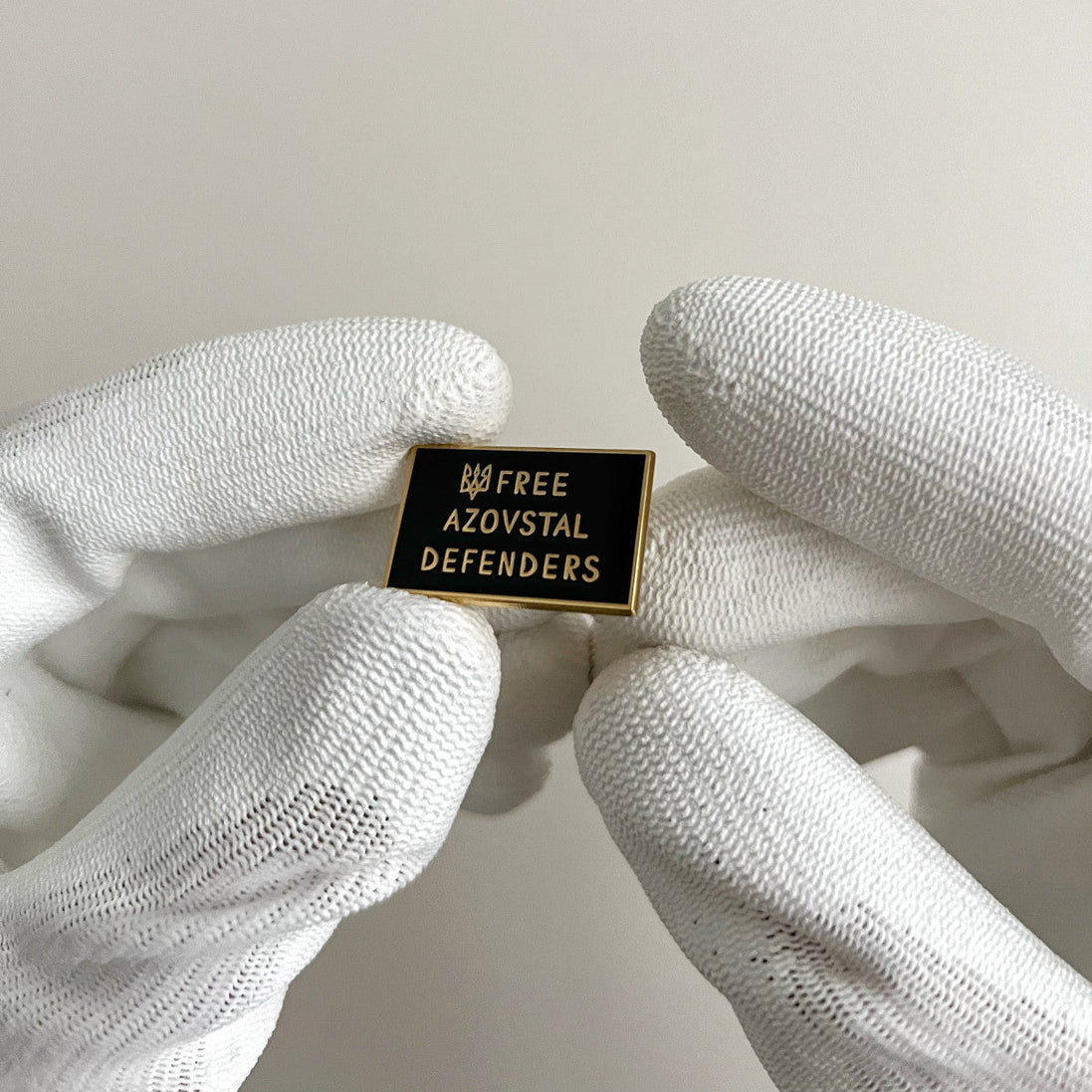 Pin with 'Free Azovstal Defenders' text held by white gloves on a light background