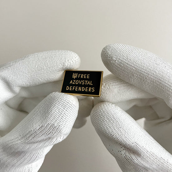 Pin with 'Free Azovstal Defenders' text held by white gloves on a light background