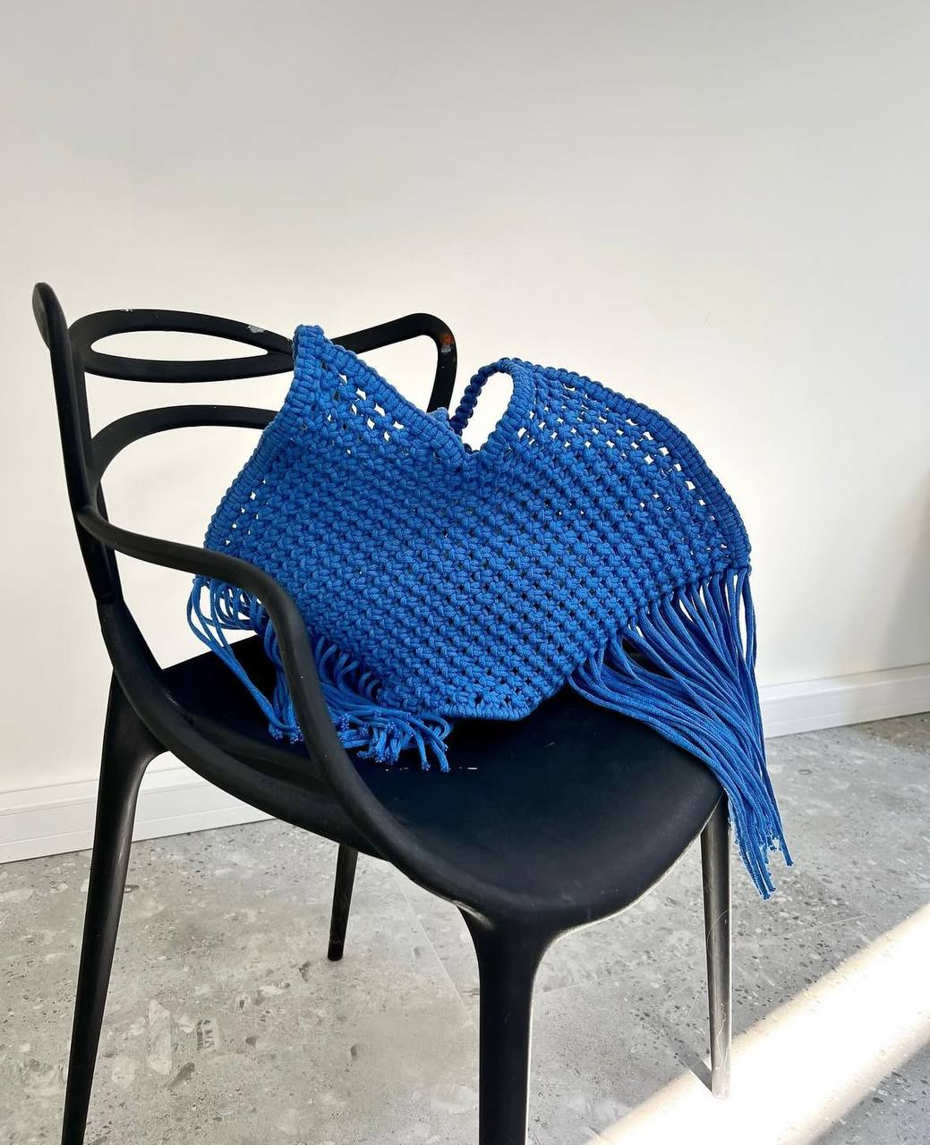 Blue woven bag with fringe on a black chair against a white wall.