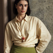 Woman wearing a cream blouse with a red flower and green skirt against a textured wall.