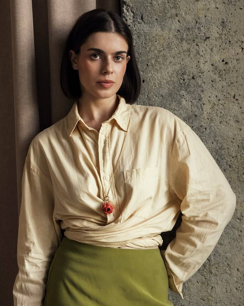 Woman wearing a cream blouse with a red flower and green skirt against a textured wall.
