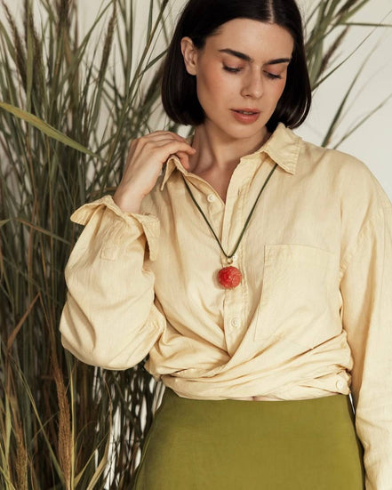 Woman wearing a beige blouse with a red pendant necklace in front of tall grass.